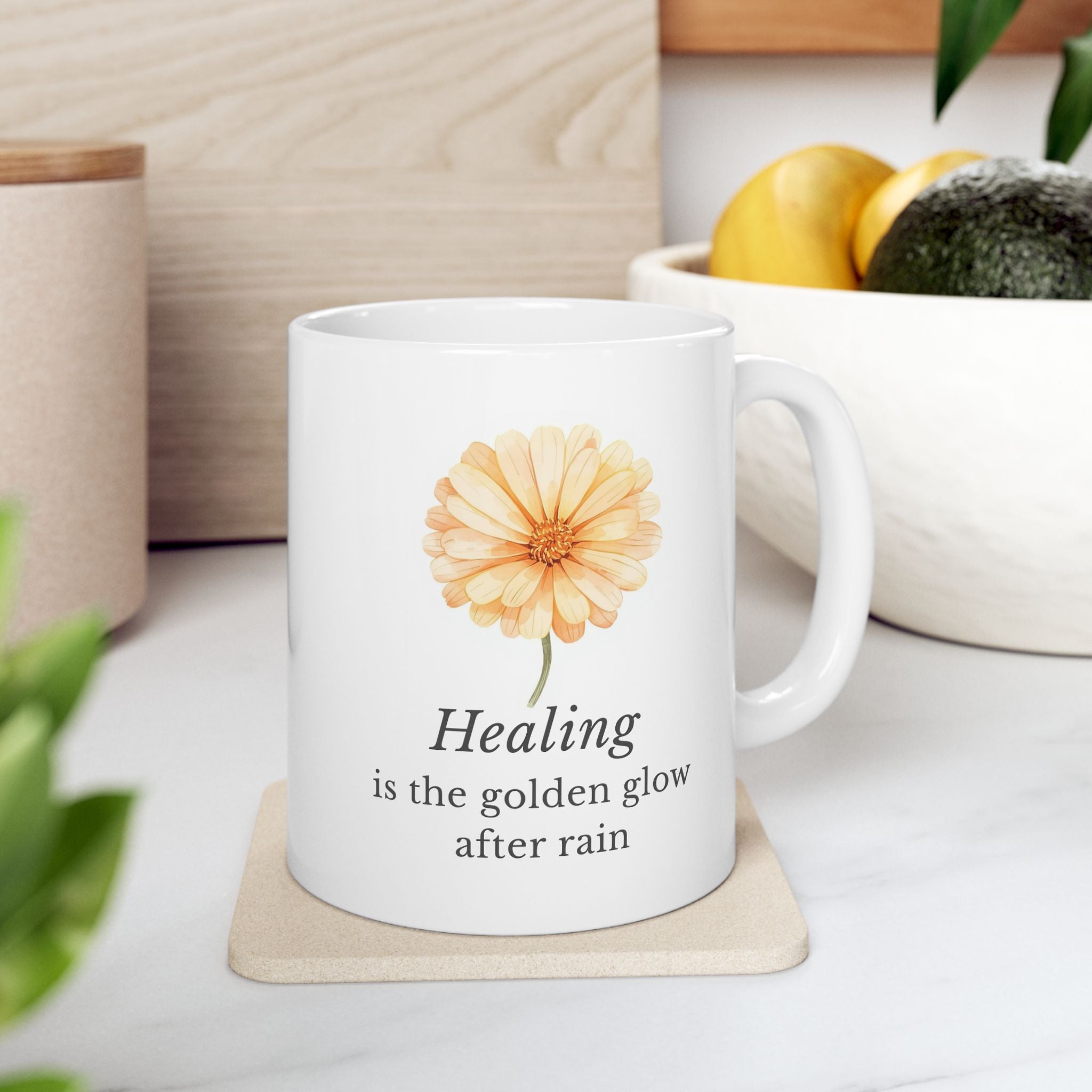 White ceramic mug with a calendula flower design and text 'Healing is the golden glow after rain' on a coaster, placed on a surface with a plant and container in the background.
