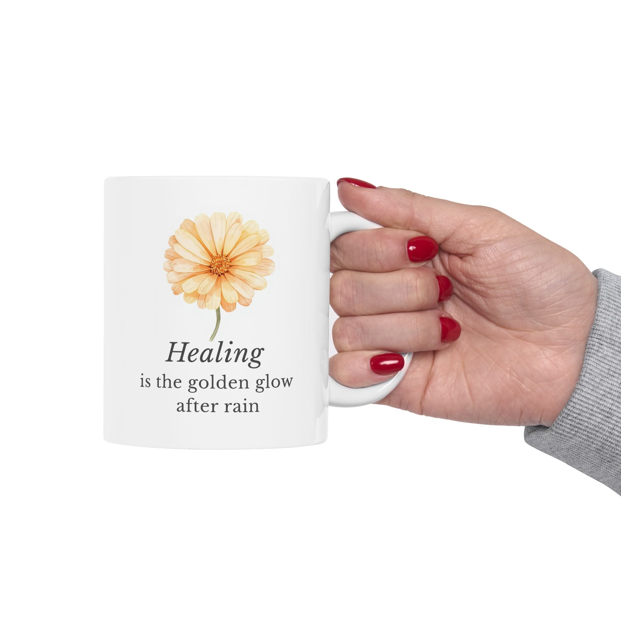 Hand holding a white ceramic mug with a calendula flower design and text 'Healing is the golden glow after rain' on a white background
