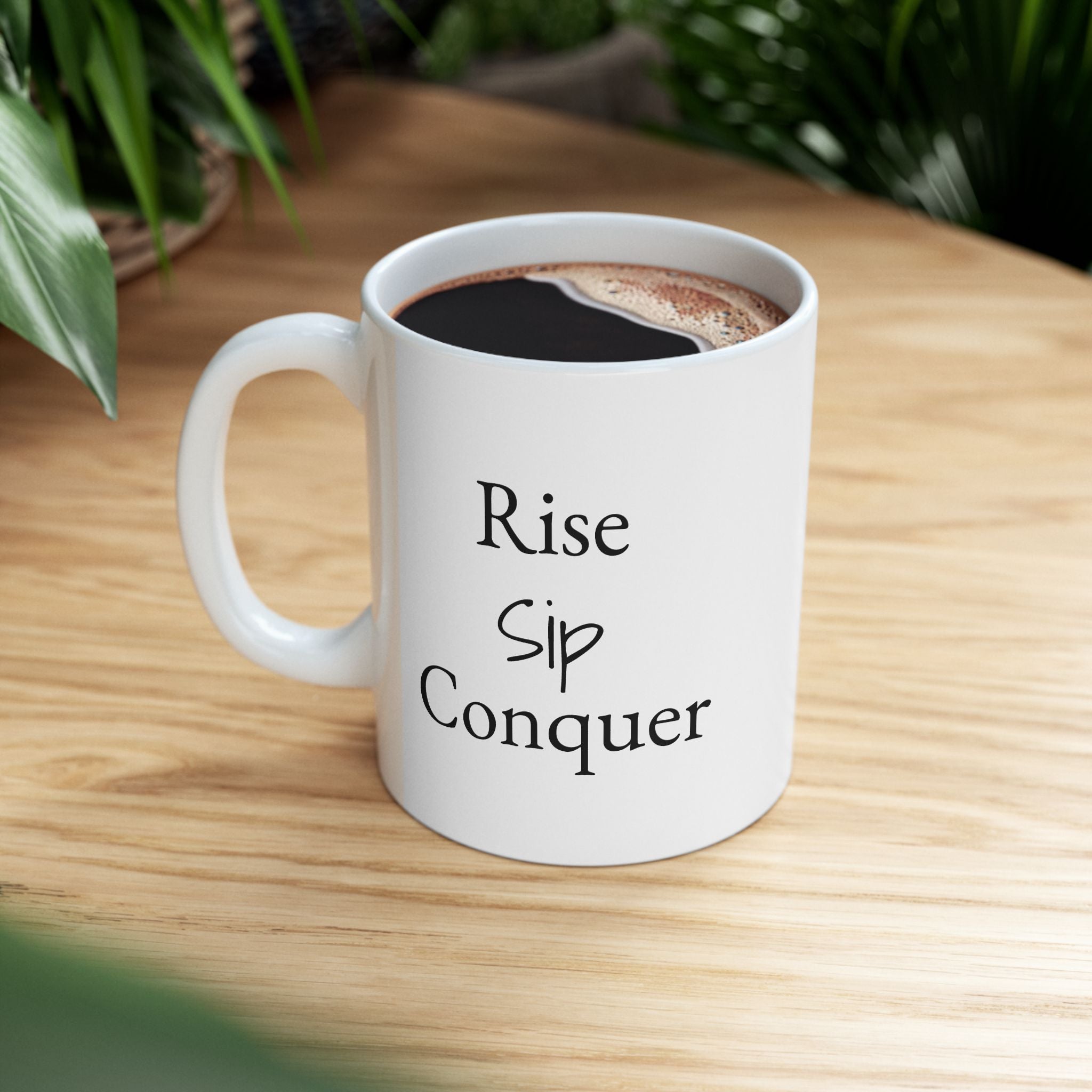 White ceramic mug with 'Rise Sip Conquer' text on a wooden table with coffee.