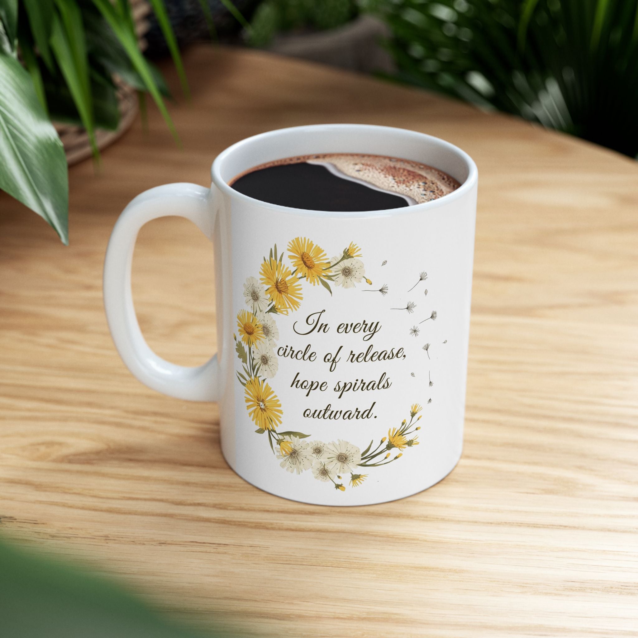 White ceramic mug with dandelion wreath design and text 'In every circle of release, hope spirals outward.' on a wooden table