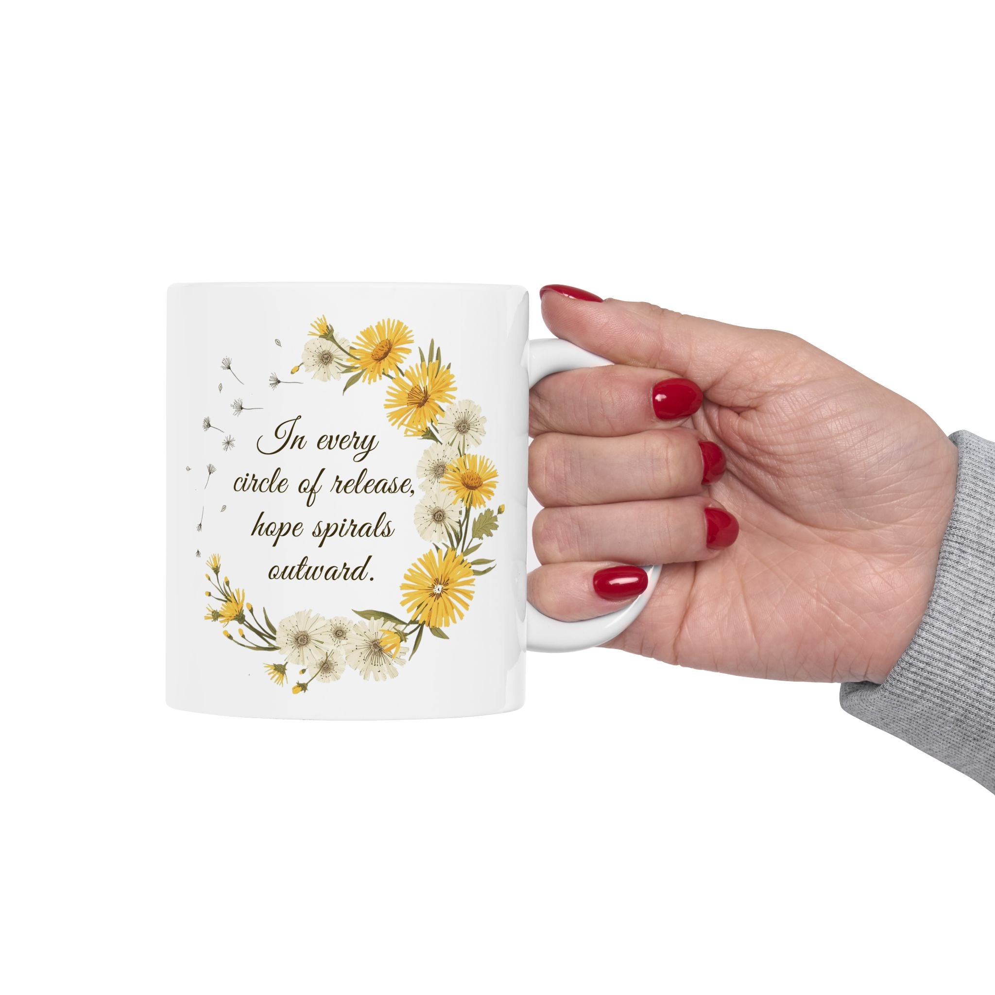 Hand holding a ceramic mug with dandelion wreath design and text 'In every circle of release, hope spirals outward.' on a white background