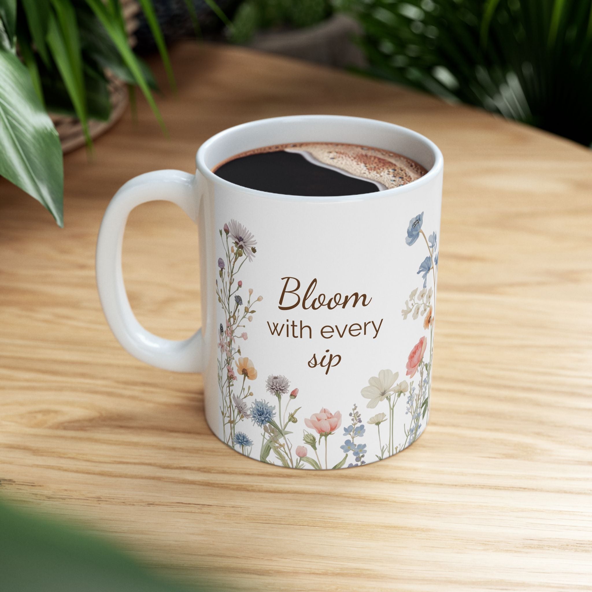 White mug with floral design and text 'Bloom with every sip' on a wooden surface.