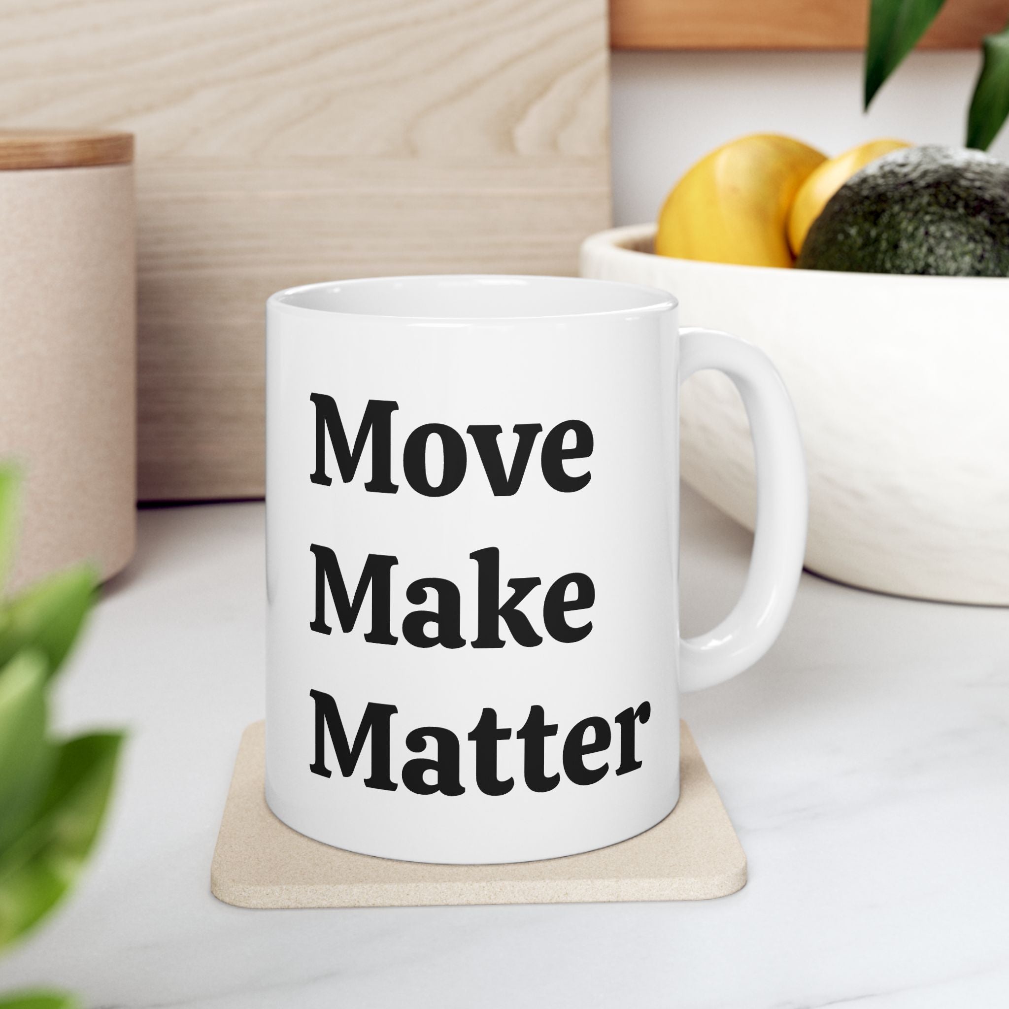White mug with 'Move Make Matter' text on a wooden coaster, surrounded by plants and a bowl of fruit.